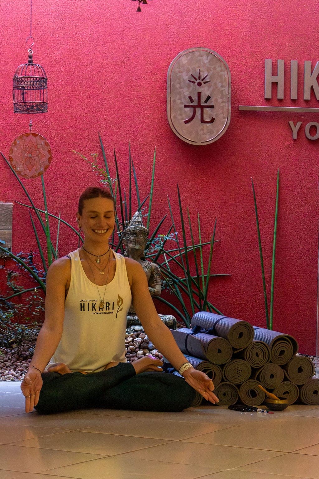 Victória Bonocielli sorrindo, sentada de pernas cruzadas de regata HIKARI diante da parede rosa com o letreiro 光 HIKARI YOGA.