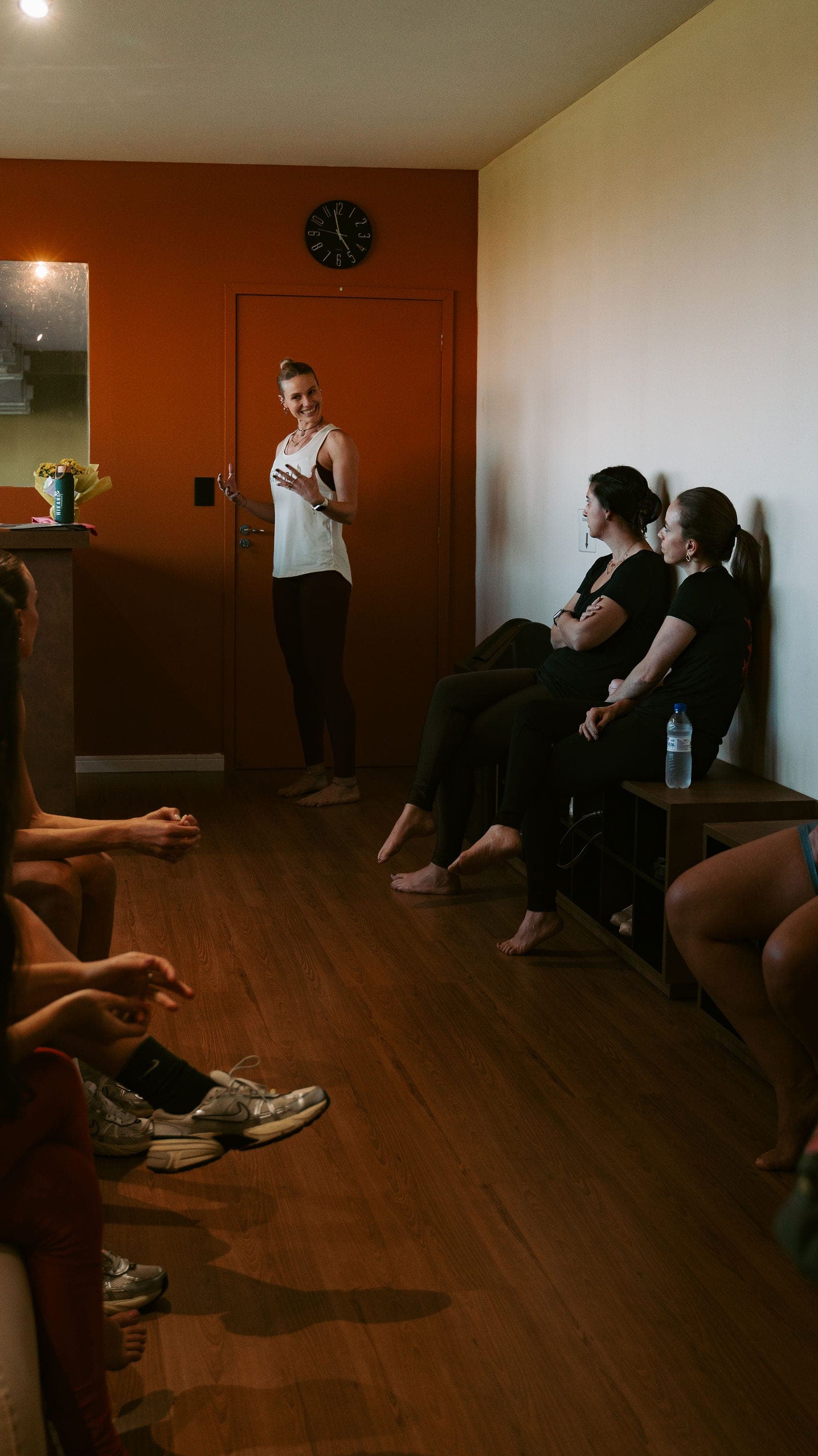Professora do Hikari de pé conversando com alunas sentadas encostadas na parede, antes da aula de hot yoga.