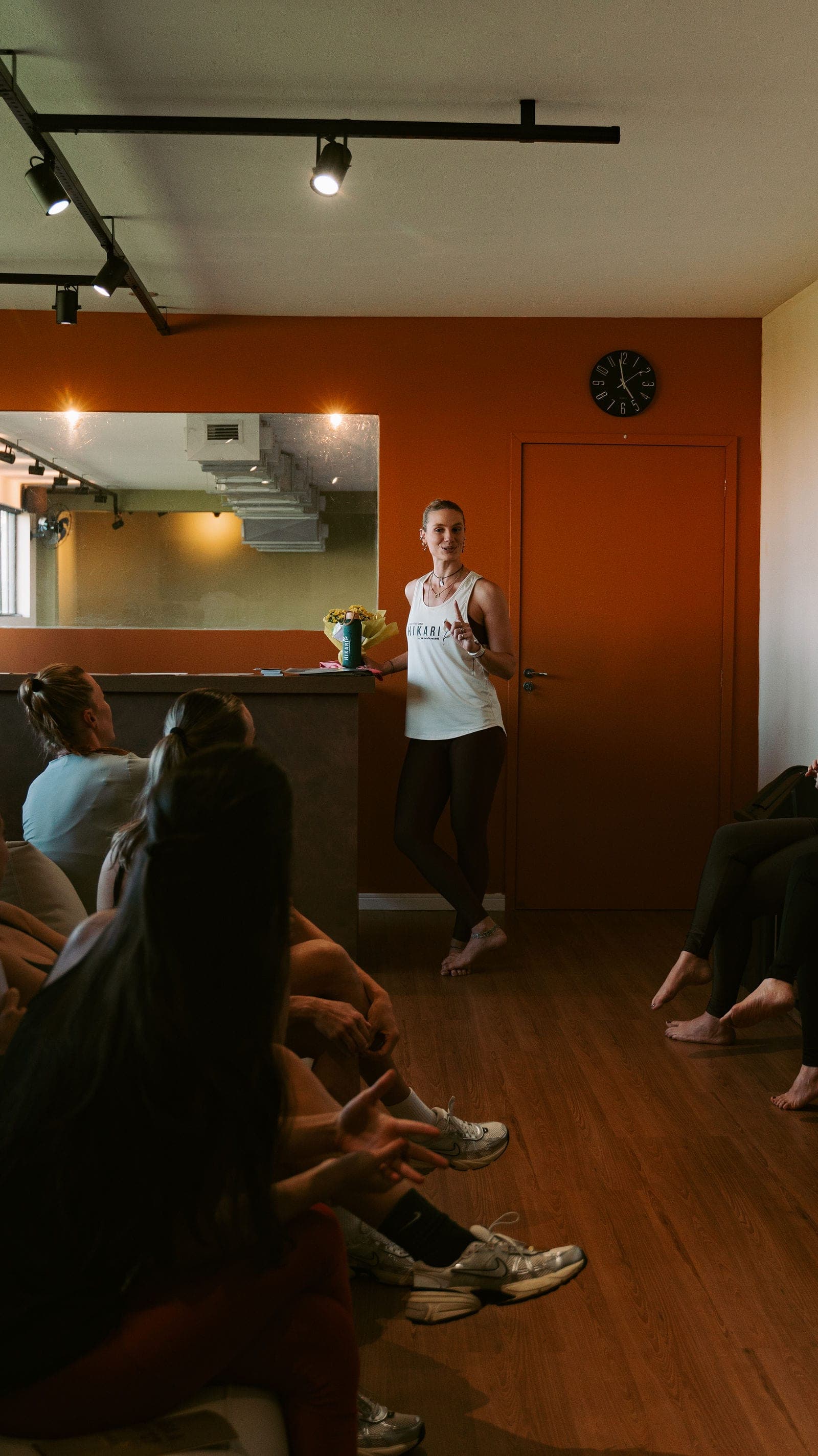 Professora do Hikari conduzindo a aula de hot yoga diante da turma sentada de costas, na sala de parede terracota.