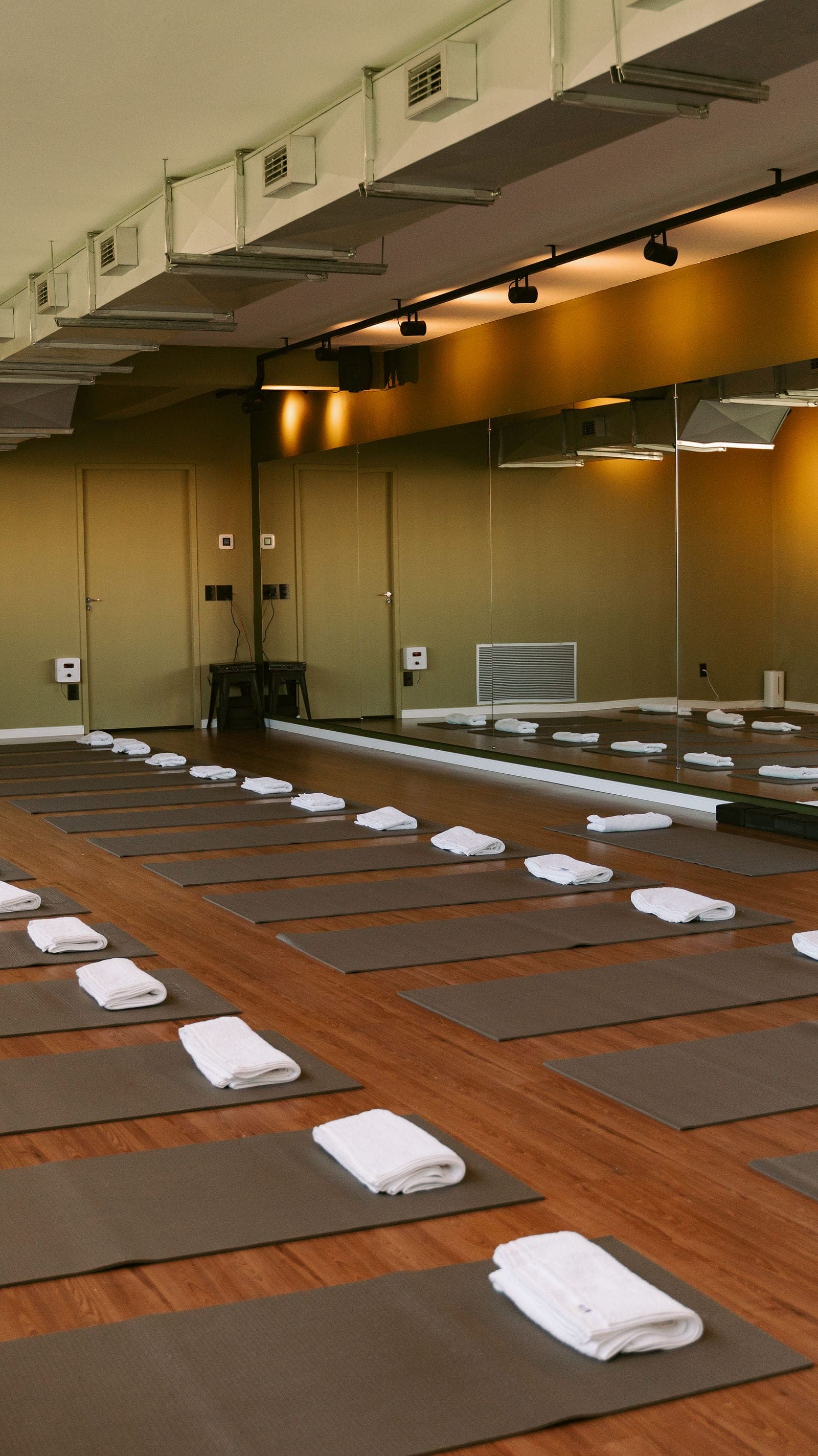 Sala de hot yoga do Hikari montada para a aula, vista pela parede espelhada, com tapetes e toalhas alinhados.