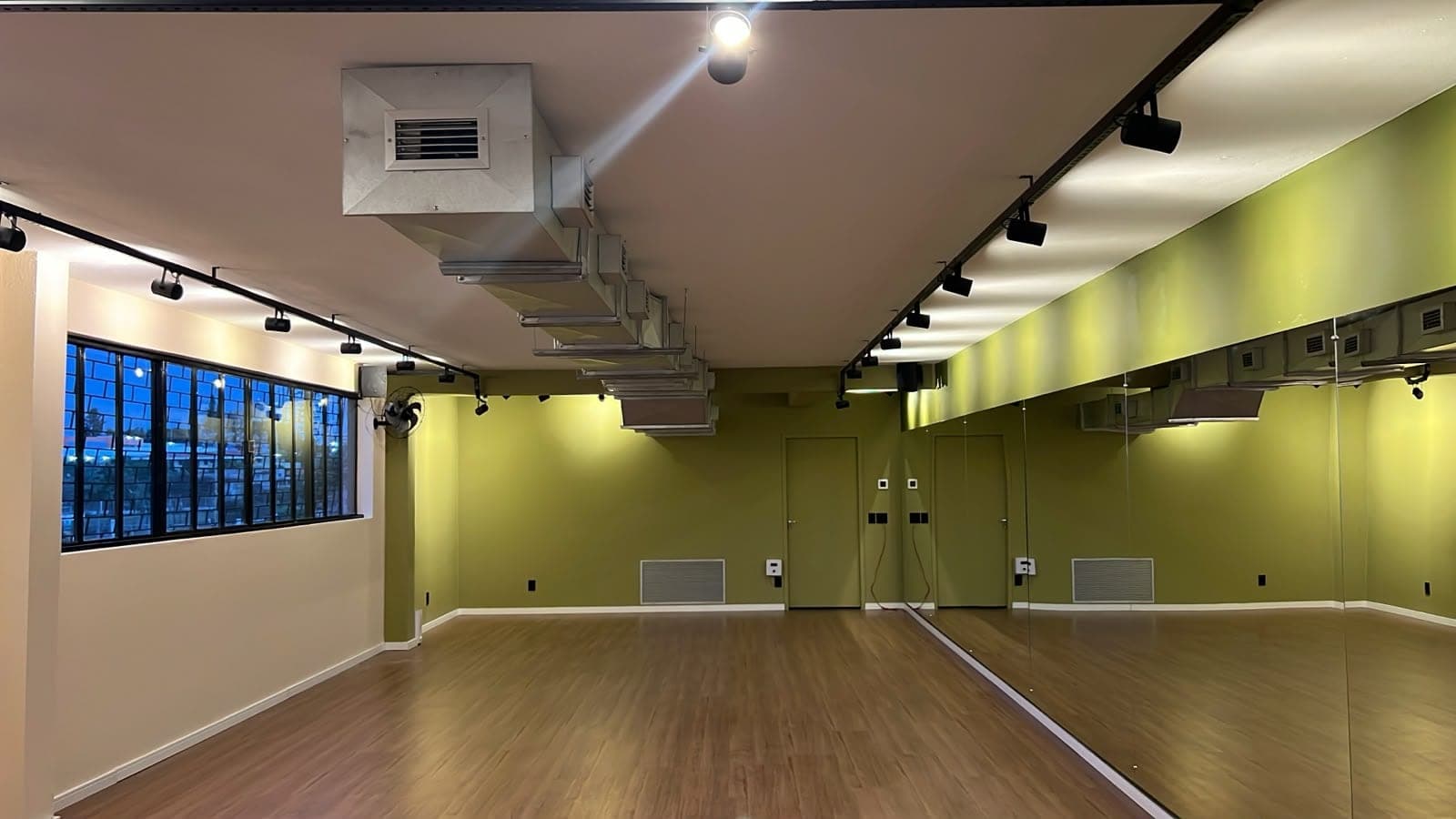 Sala de hot yoga do Hikari em perspectiva pela parede espelhada, com janelas ao entardecer e piso de madeira.